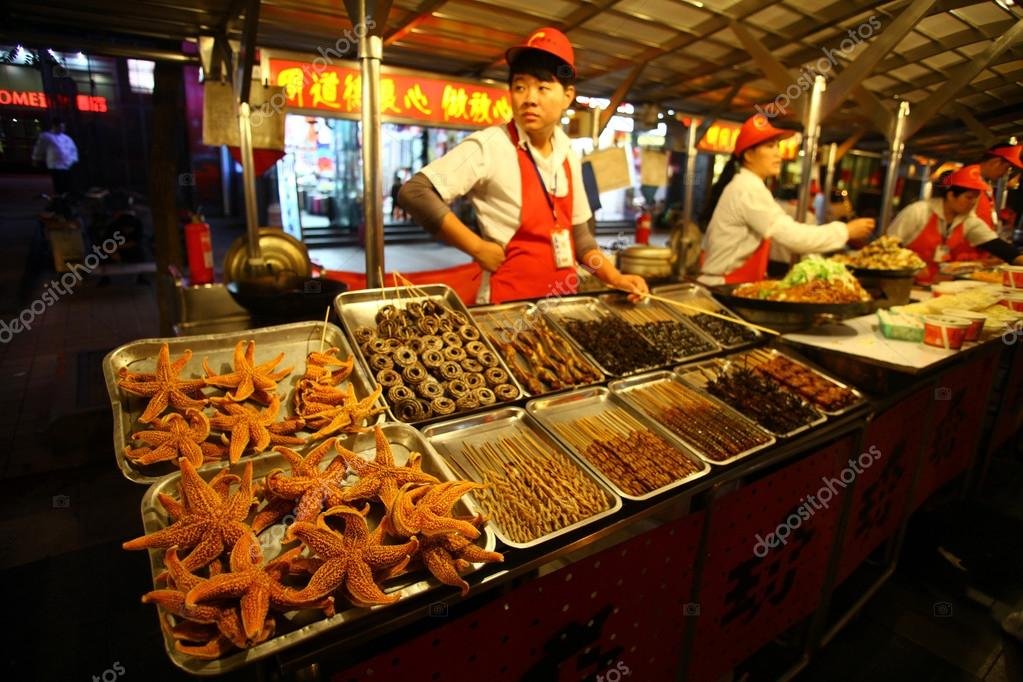 A vendor at Donghuamen Yeshi night market in Beijing presenting trays of exotic snacks, including starfish and scorpions on skewers, for adventurous visitors.